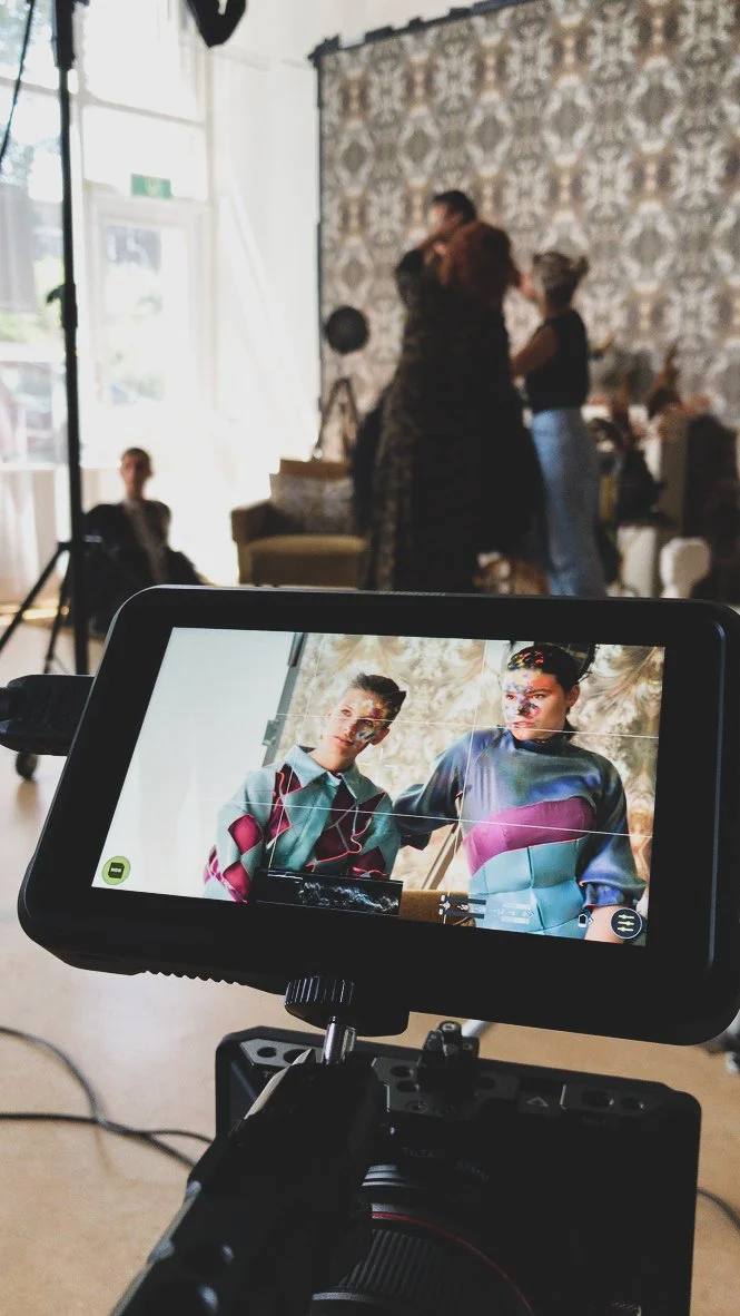 A woman with short gray hair and a black shirt takes a photo of two young women with braids, wearing colorful tops, in a room with patterned wallpaper. The photo is being taken with a camera on a tripod, showing the scene on its screen.