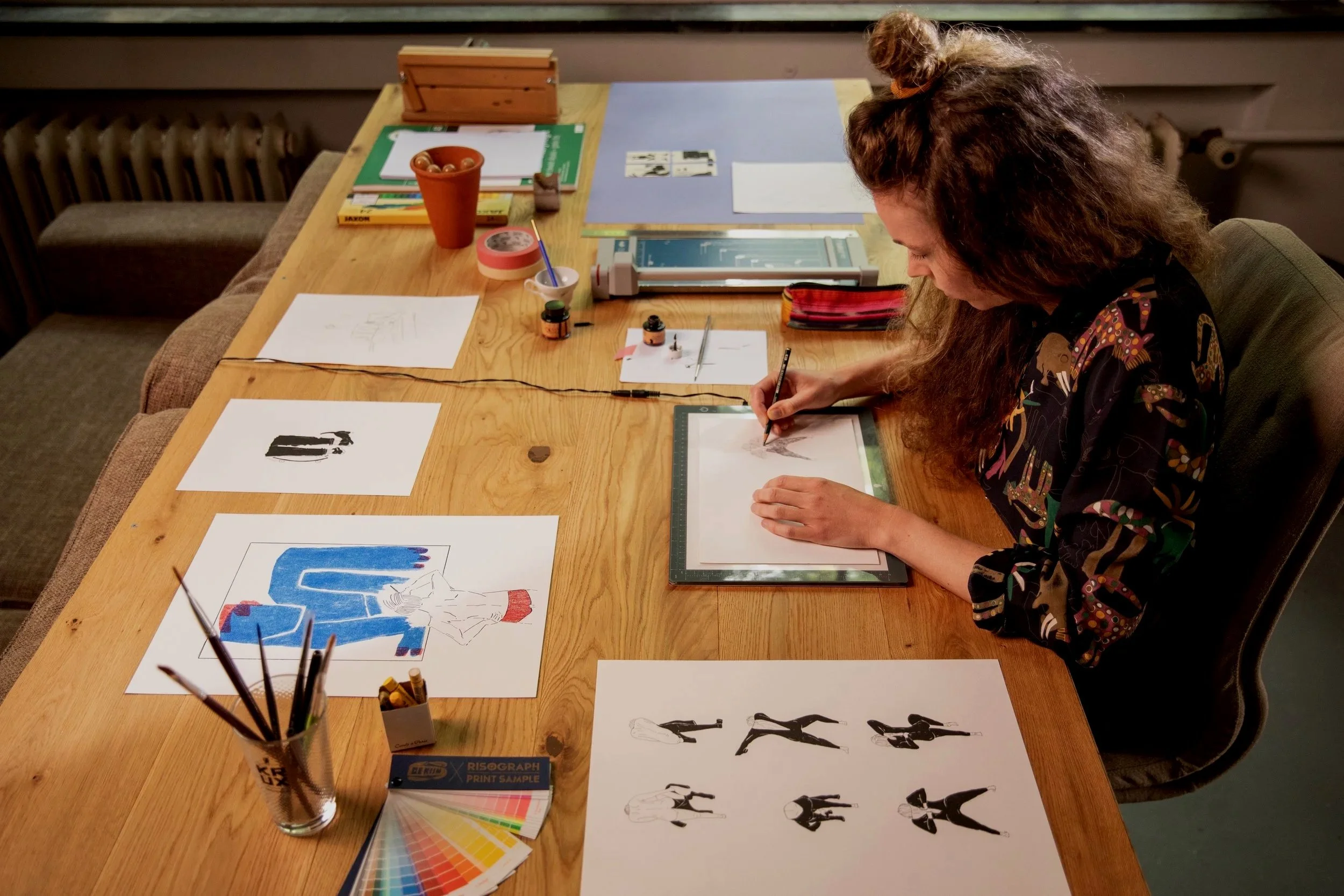 A woman with long curly hair and glasses drawing on a tablet at a wooden table that has various art supplies including ink bottles, brushes, sketches, colored artwork, and a color palette sample sheet.