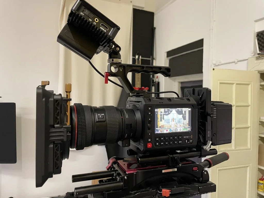 Camera setup with a large professional video camera on a tripod, with an external monitor attached, in a room with shelves and a closed door in the background.