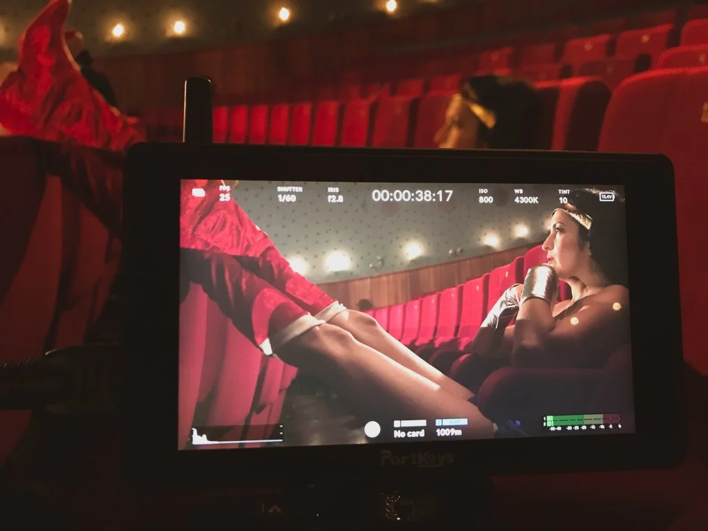 A woman wearing a gold headband, black gloves, and silver bracelets sits in a red theater seat, looking stage right with a contemplative expression. The background shows more red theater seats and a wall with ceiling lights.