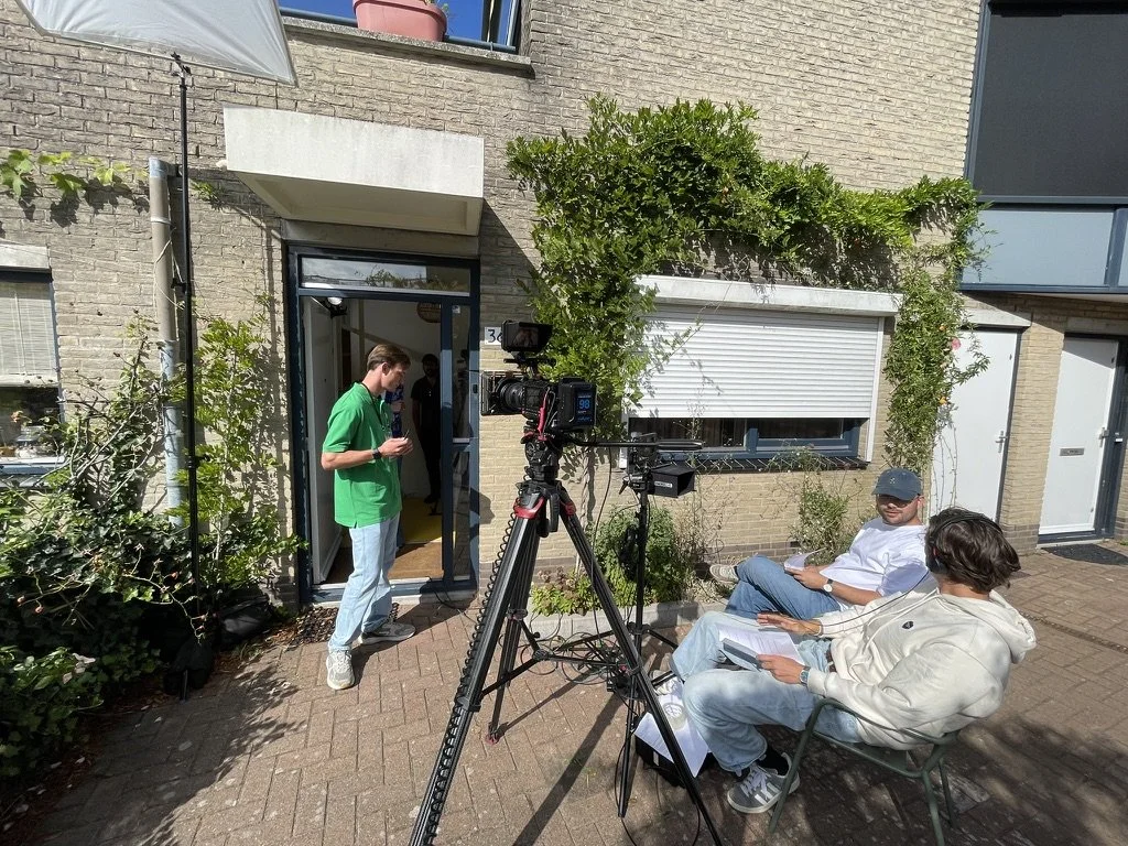 Two people sitting outdoors with notebooks, facing a camera on a tripod; one in a white hoodie and sunglasses, the other in a white shirt and cap; a young man in a green shirt standing at the door.