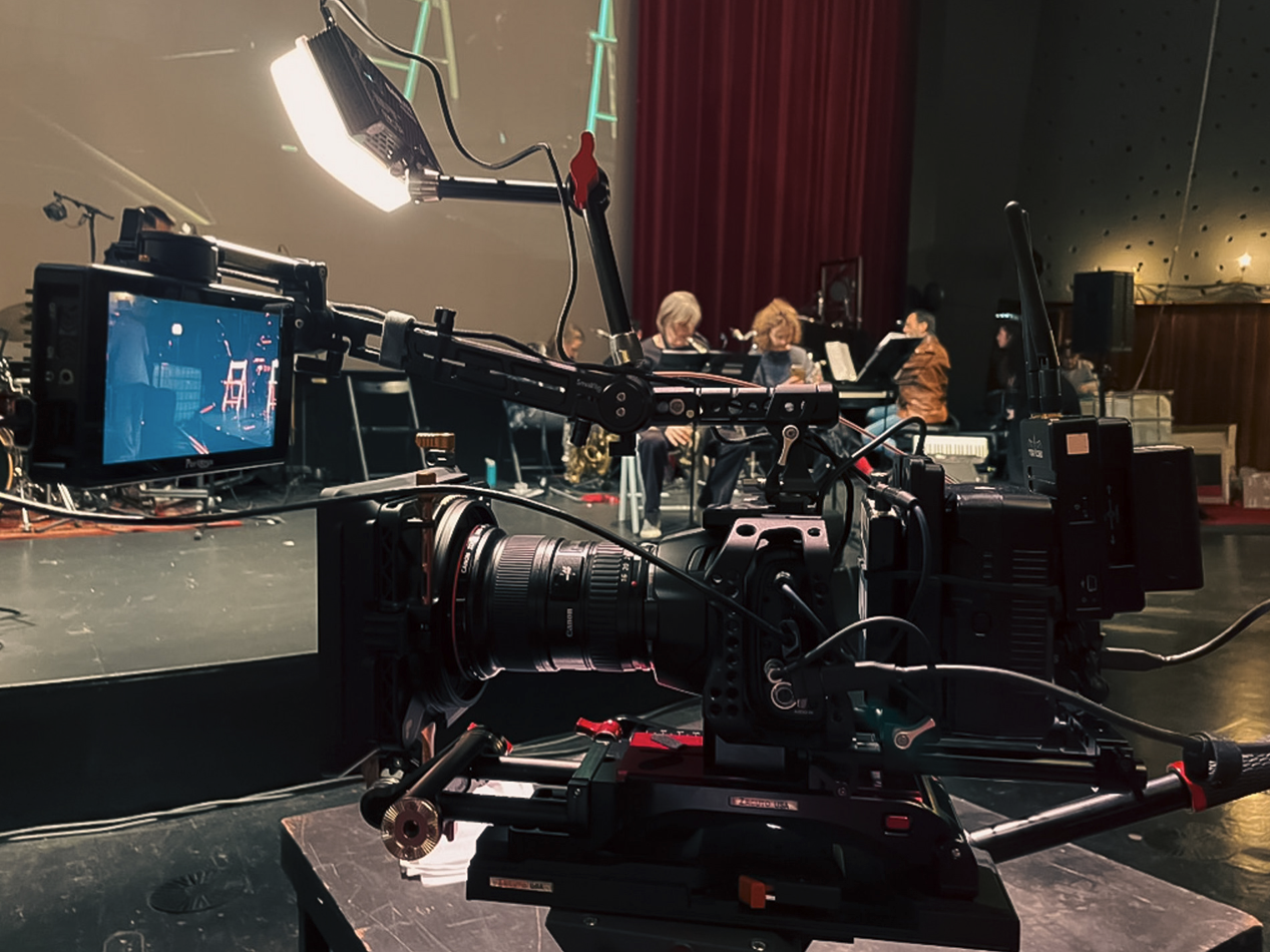 Camera gericht op een orkest dat oefent op een podium, met muzikanten die bladmuziek lezen.