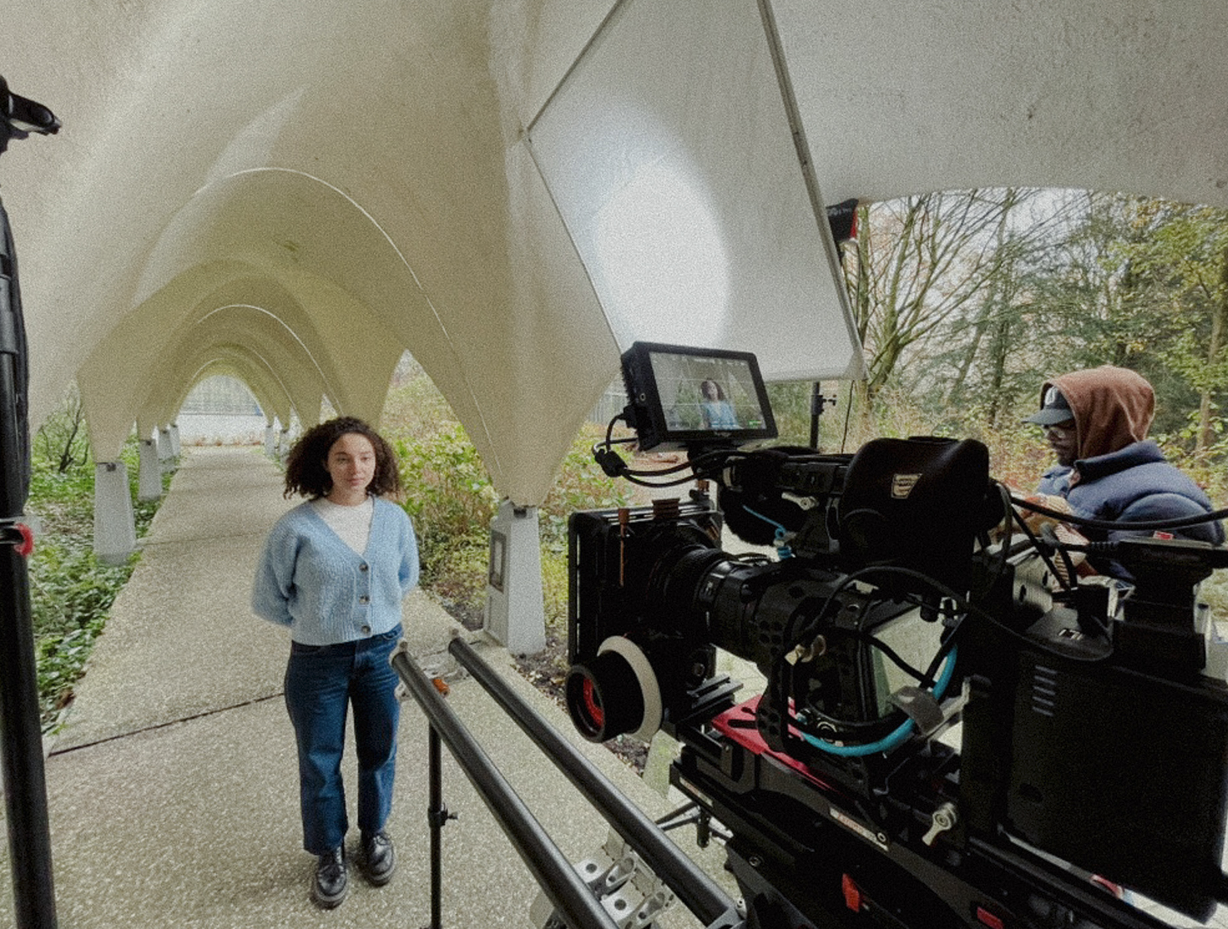 Een vrouw in een blauwe trui en spijkerbroek die zich voordoet voor een filmcamera onder een witte, grafische structuur in een bosrijke omgeving.