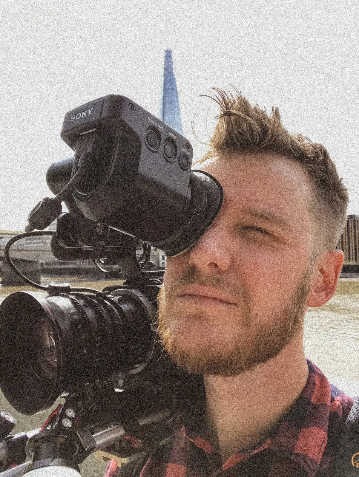 A man with a beard and a checkered shirt taking a photo with a professional video camera on his shoulder, standing outdoors near water with city buildings in the background.