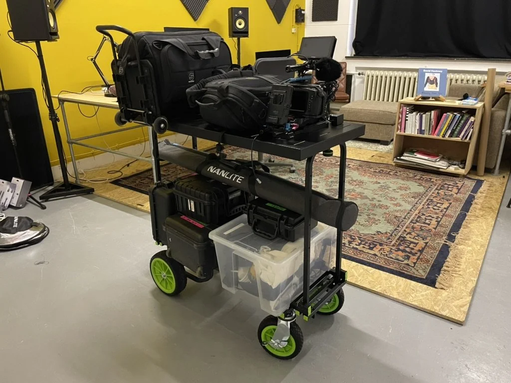 Mobile cart with camera equipment, bags, and accessories in a music or recording studio with yellow walls, bookshelves, and seating area.