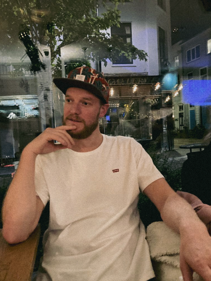 Man in wit T-shirt en een pet, zittend in een restaurant of café, met een ramenachtergrond en straatverlichting te zien.