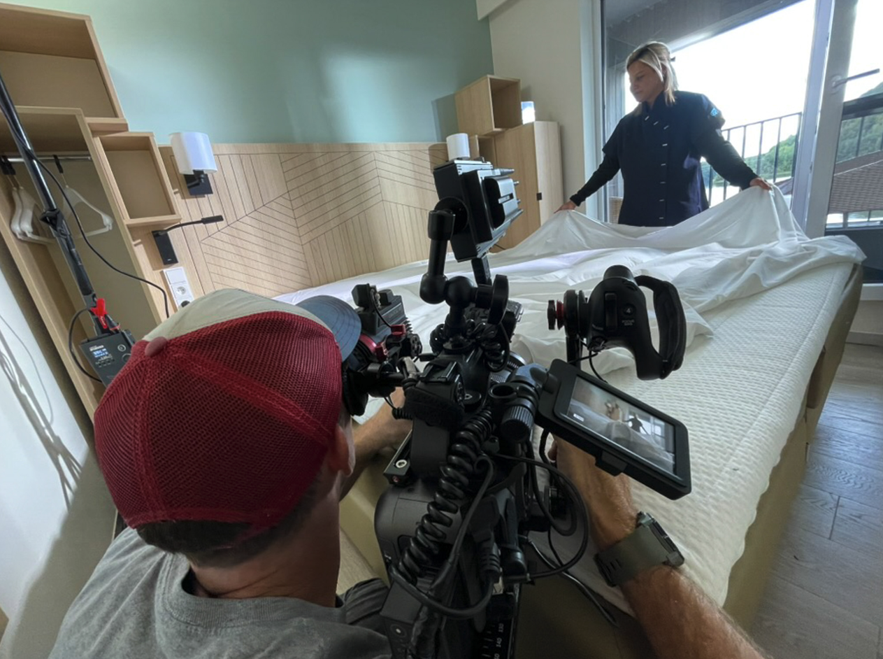 Een persoon met een rode pet en camera richt zich op een bed, terwijl een vrouw het bed opmaakt in een kamer met grote ramen en balkon. Film- of fotoshoot opnames zijn bezig.