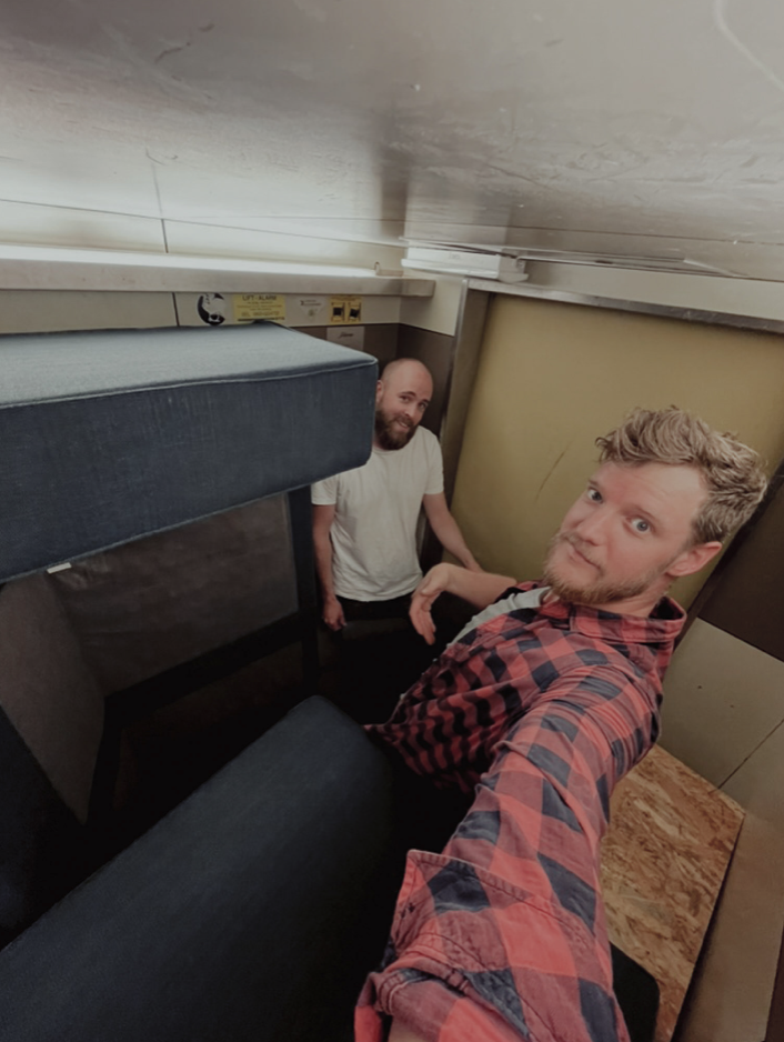 Twee mannen maken een selfie in een kleine, smalle kamer met bedden, een paar stopcontacten en een houten kast.