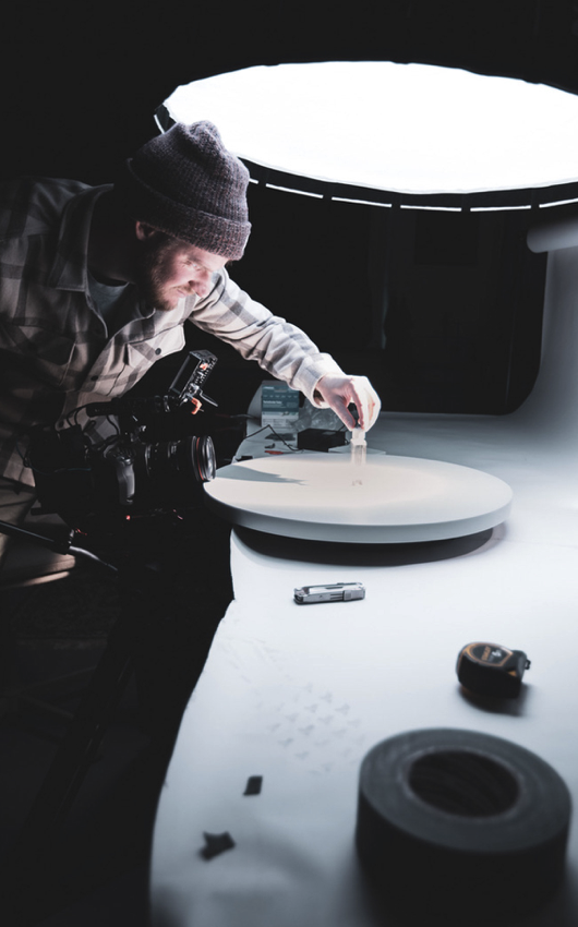 A photographer wearing a beanie and plaid shirt adjusting a small object on a circular white platform, illuminated by a large diffused light source in a dark studio.
