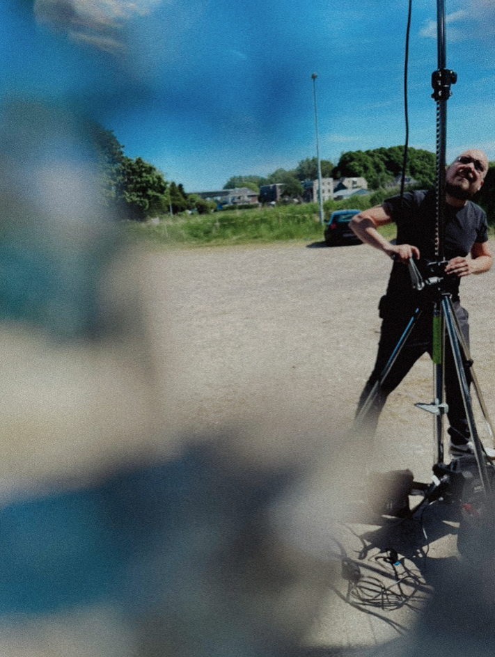 Man staat met een camera op een statief in een open veld op een zonnige dag, met huizen en auto's op de achtergrond.