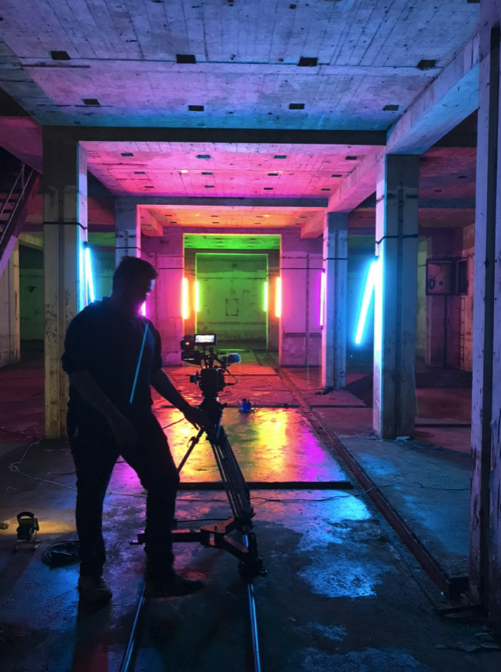 Een persoon bedient een camerastatief in een ruimte met neonverlichting in verschillende kleuren, waaronder blauw, geel, groen, roze en oranje.