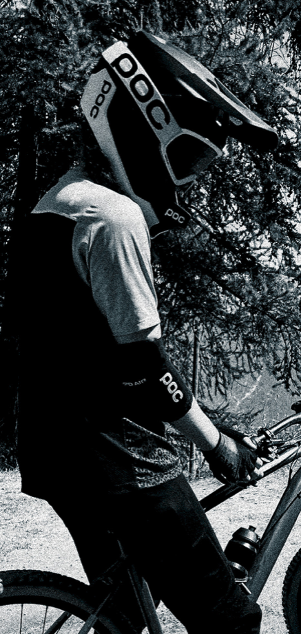 Persoon in een mountainbikoutfit met een POC helm en gordels, die een mountainbike vasthoudt, in een bosrijke omgeving.