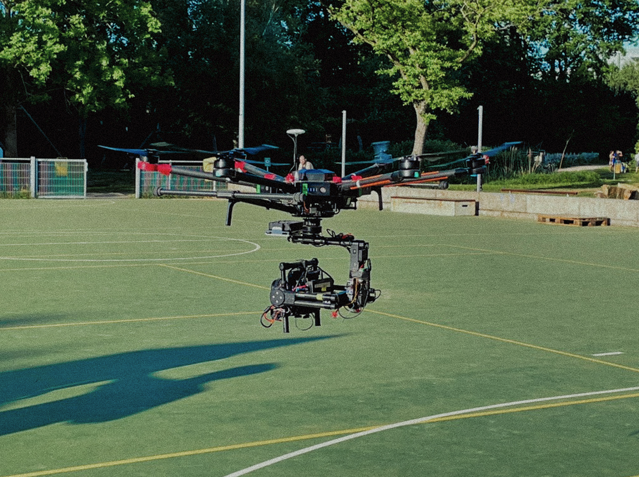 Drones vliegen boven een sportveld terwijl mensen in de achtergrond wandelen en genieten van het weer.
