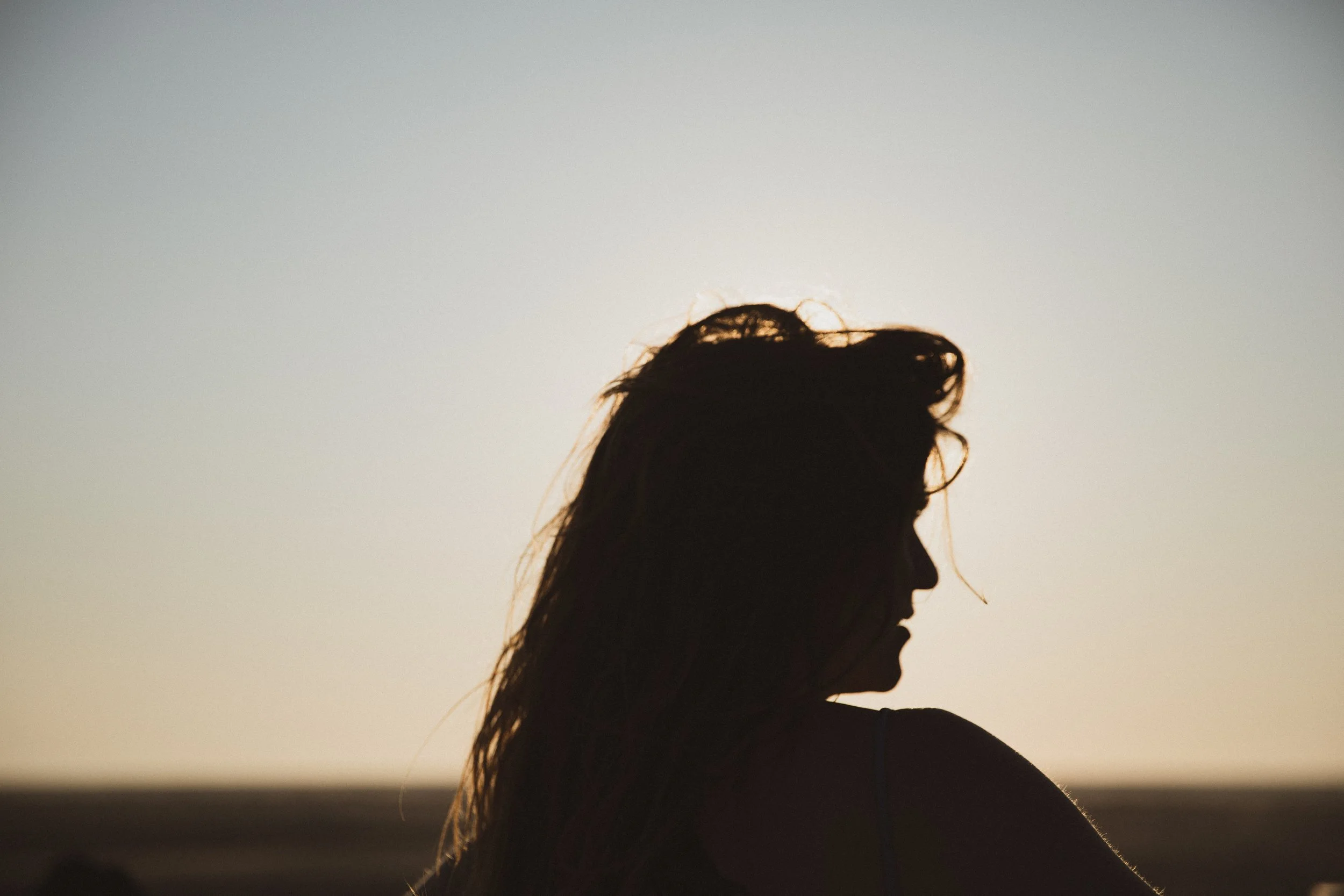 Silhouette of a woman with long hair looking down against a beach at sunset.