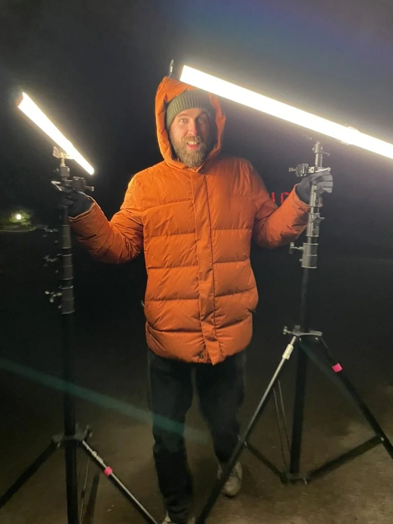 A man dressed in a brown winter jacket and gray beanie stands outdoors at night, holding two LED light panels on tripods.