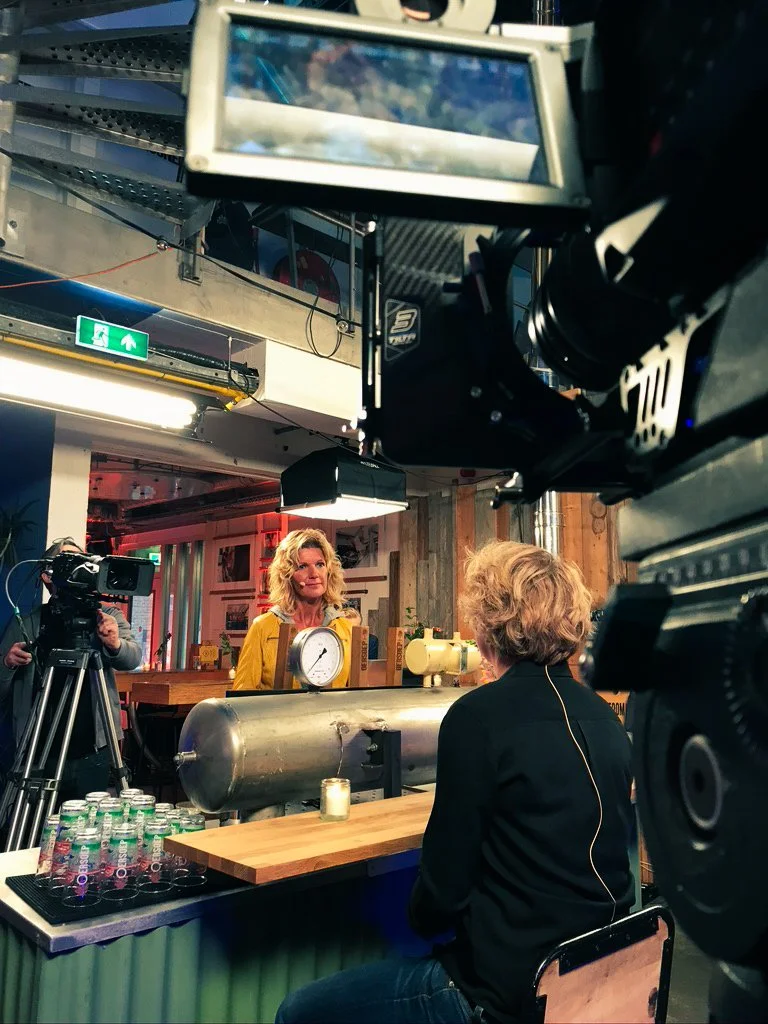 A woman in a yellow jacket being interviewed in a recording studio with cameras and lighting equipment, a large metal tank and bottles of water on a table in front.