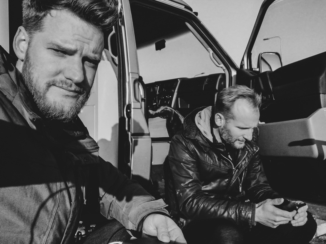 Twee mannen zitten naast een grote truck, een van hen kijkt naar de camera terwijl de ander naar een telefoon kijkt, beiden dragen leren jassen en zitten op de grond.