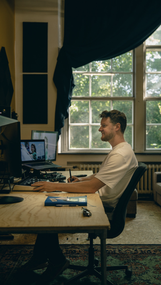 A man sitting at a desk in a home office, using a computer with two monitors, with a large window and blue curtains behind him.