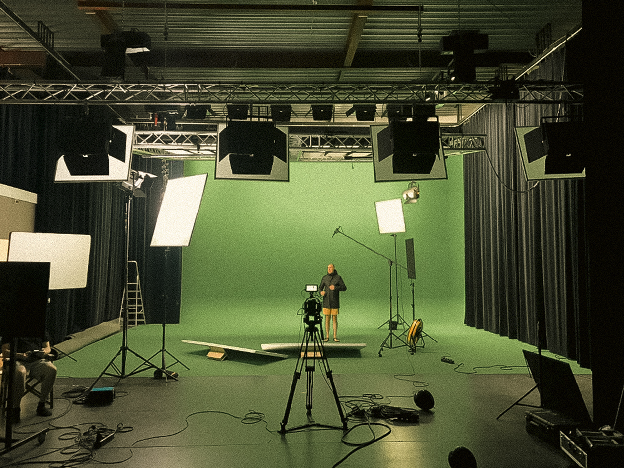 Filmstudio met een groen scherm, camera, verlichtingsapparatuur en een persoon die zich voorbereidt voor opnames.
