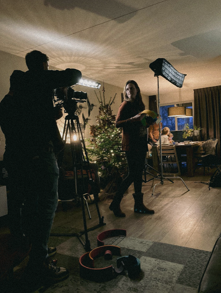Een vrouw staat in een woonkamer met een kerstboom, terwijl een cameraman en lichtapparatuur haar filmen voor een video-opname.