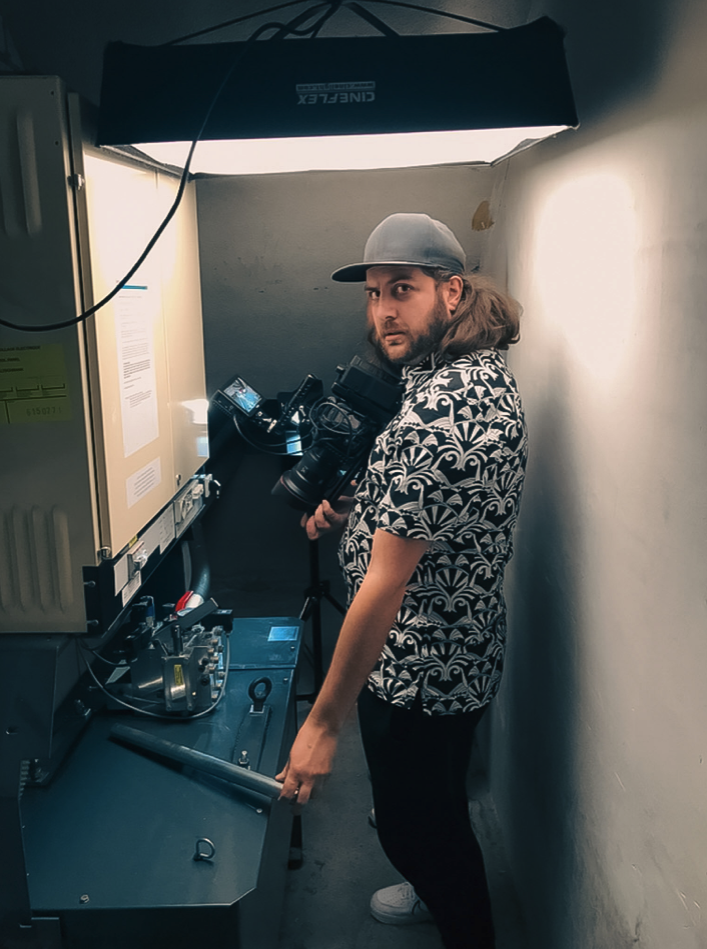 Mannelijke persoon met lange haar en hoed die een camera en apparatuur bedient in een kleine, koele ruimte met een bovenverlichting.