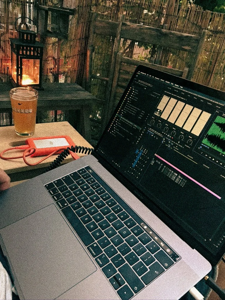 Laptop computer open to video editing software on a table outdoors with a glass of beer and an orange phone nearby.
