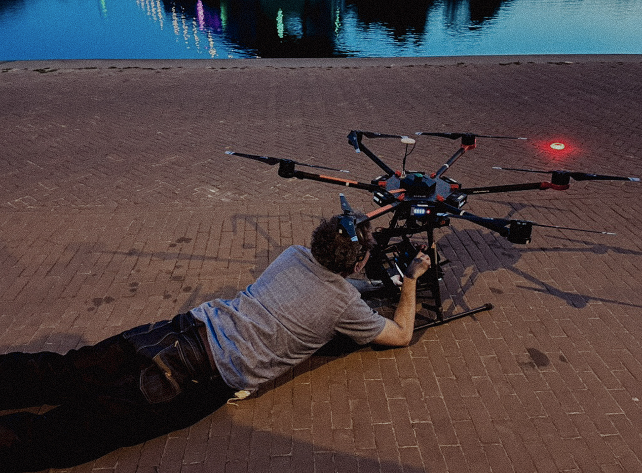 Persoon ligt op de grond en bedient een grote drone op een brickstenen straat, met water en kleurrijke lichten op de achtergrond.