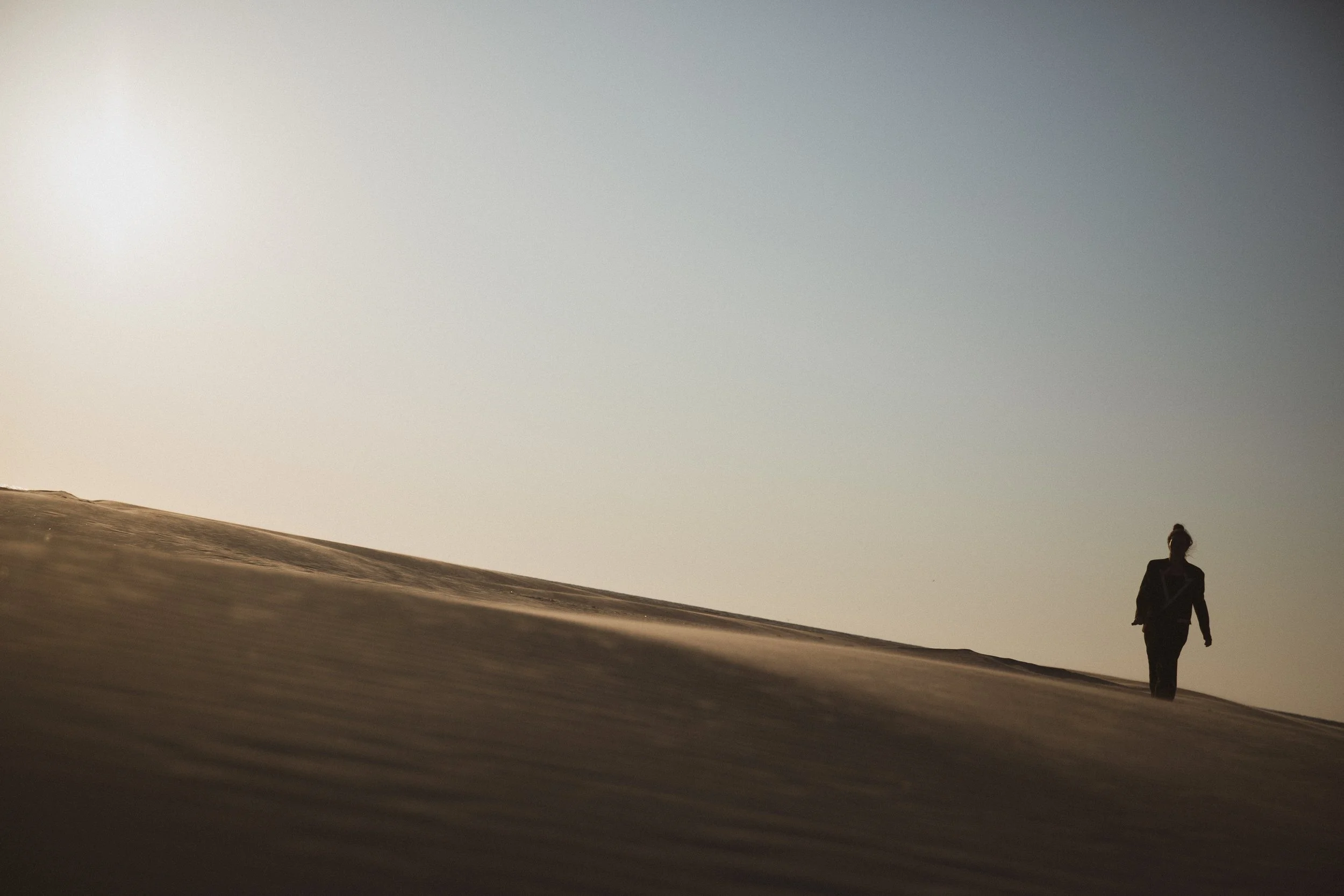 A person walking alone on sand dunes in a desert during sunset or sunrise, with a clear sky in the background.