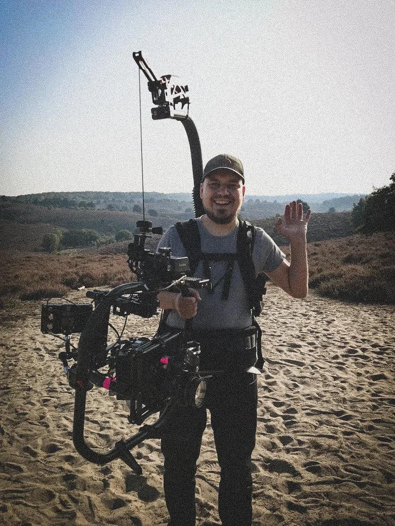 A man in outdoor gear, holding a professional camera stabilizer rig, standing on a sandy trail in a rural area, smiling and waving, with hills and trees in the background.