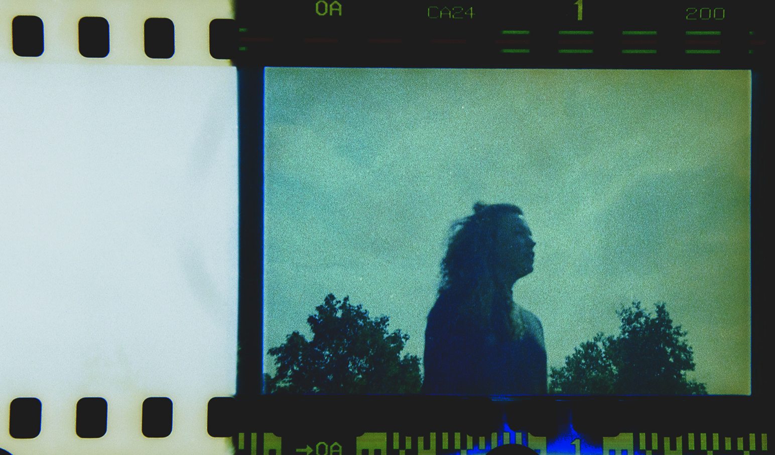 Een filmrol met een afbeelding van een silhouet van een vrouw met lang haar, zichtbaar tegen een groene achtergrond met bomen, gefotografeerd op een reflectief oppervlak met andere filmranddetails.