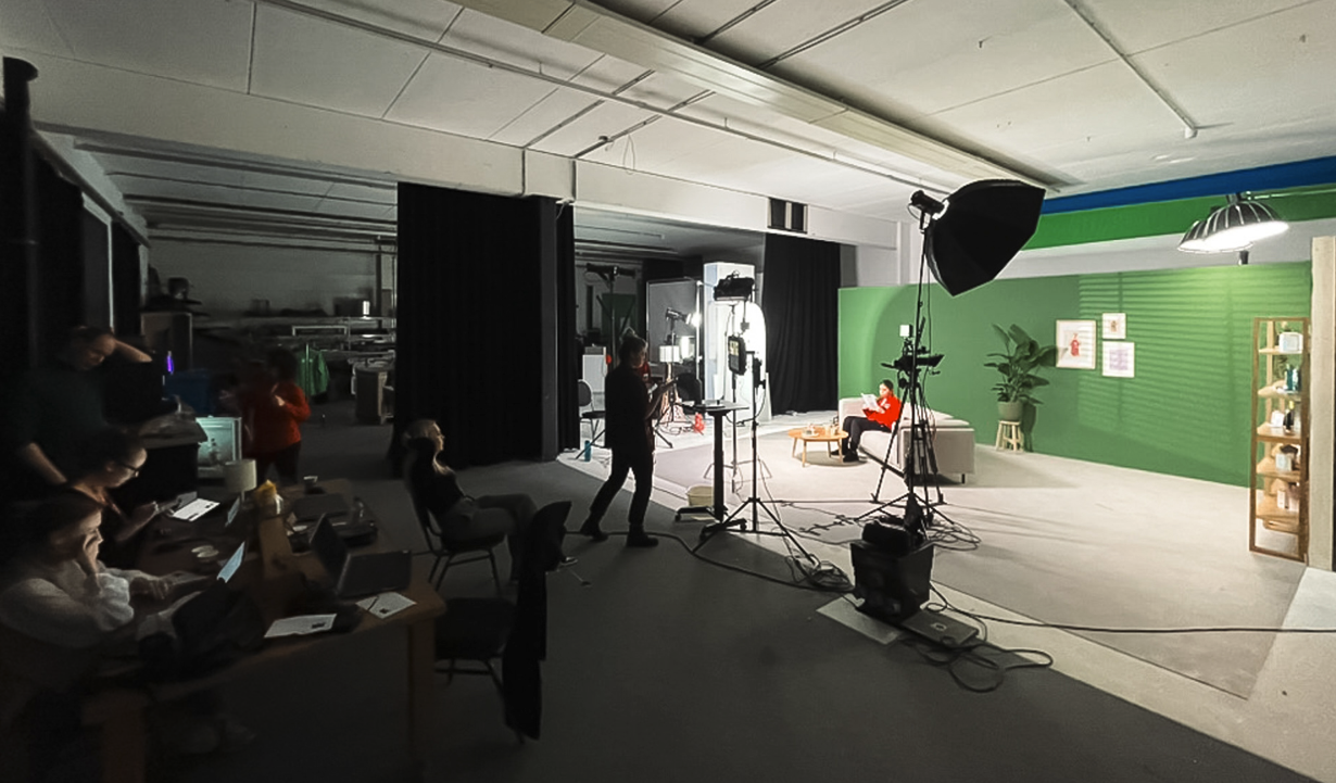 Een opnamestudio met een groen achtergrond, camera's, verlichting en een persoon die op een sofa leest, klaar voor een opname of fotoshoot.