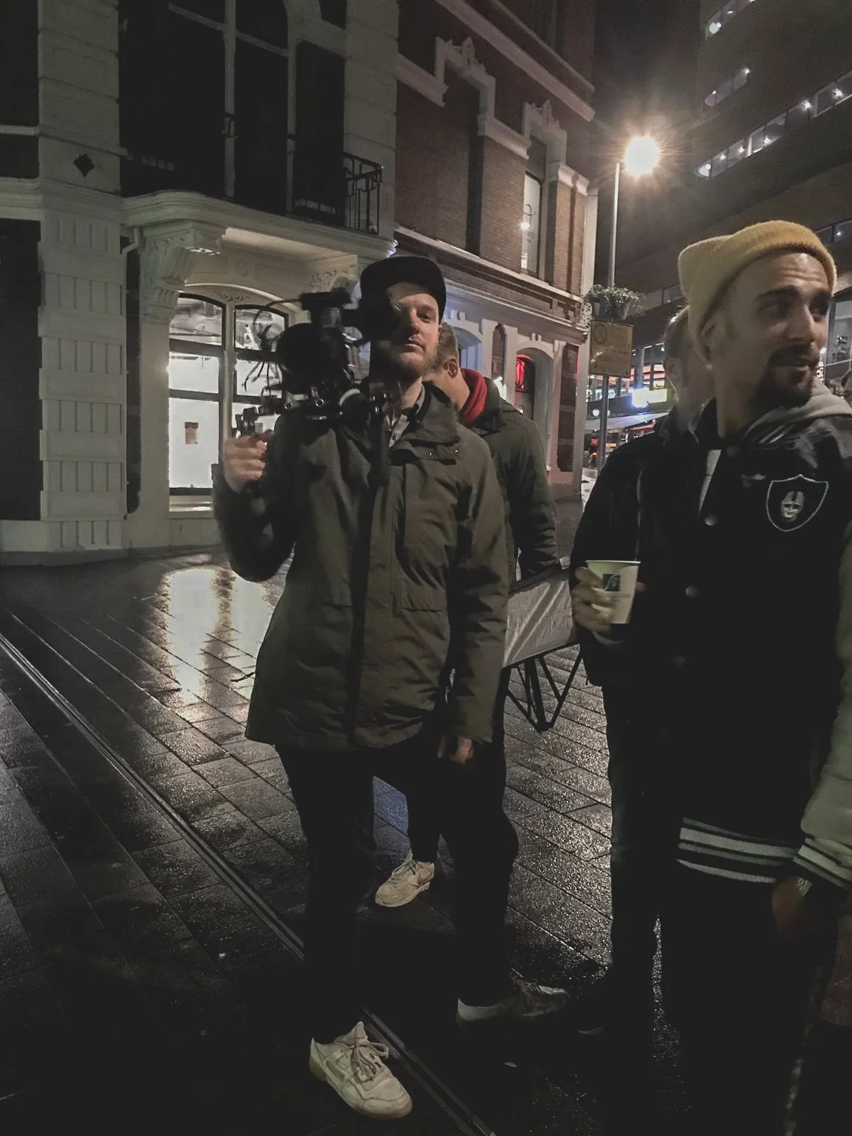 A group of men standing on a wet, dark street at night, with one man holding a camera and wearing a black beanie, and another man holding a cup, with a historic-style building and streetlights in the background.