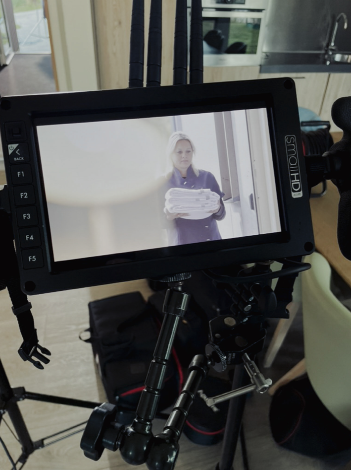 Beeld van een filmcamera gericht op een vrouw die een hoop papieren vasthoudt, binnen in een kamer met een keuken op de achtergrond.