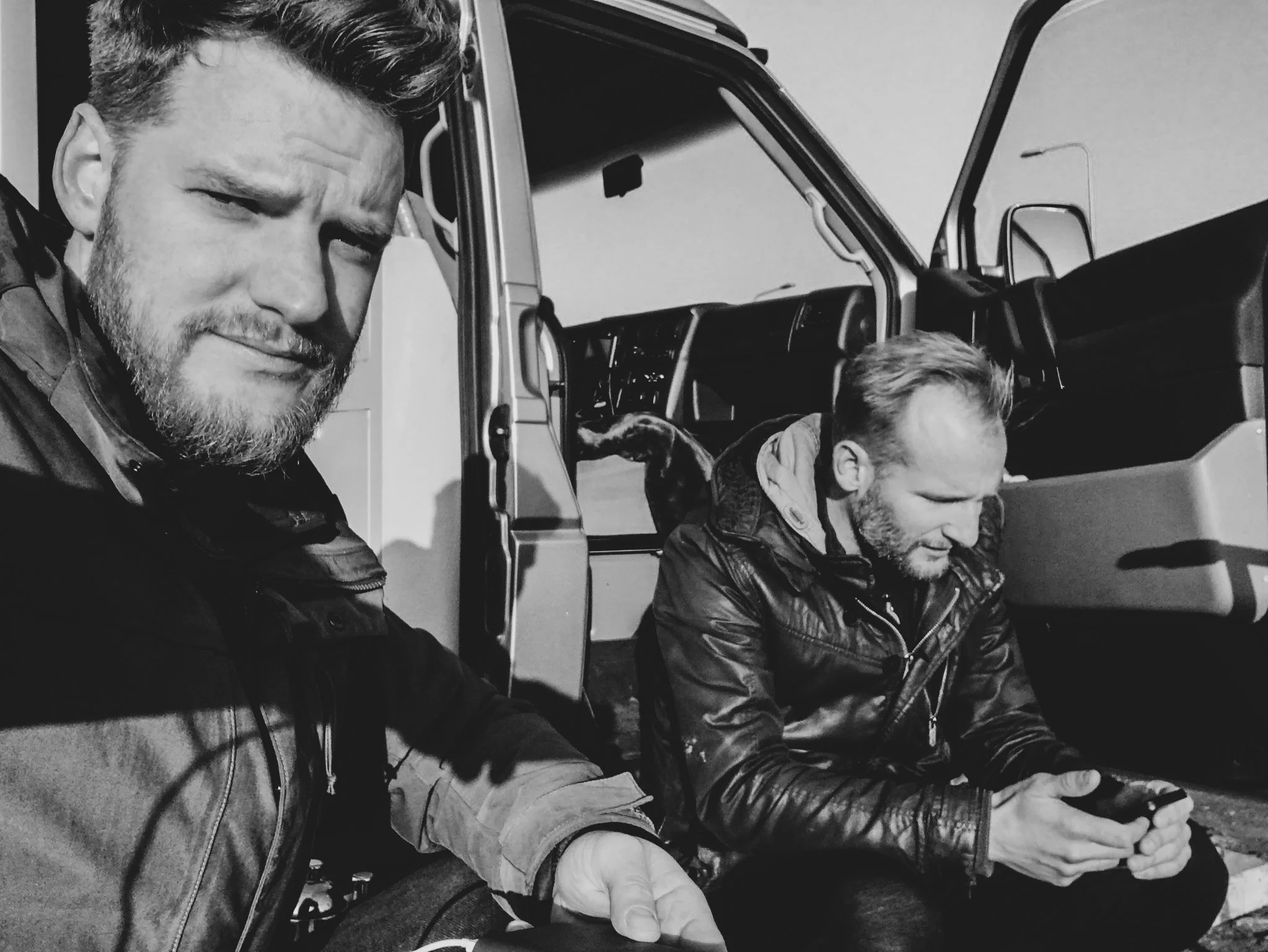 Two men sitting on the ground beside a large truck, one is taking a selfie, both are looking at their phones, outdoor setting, black and white photo.