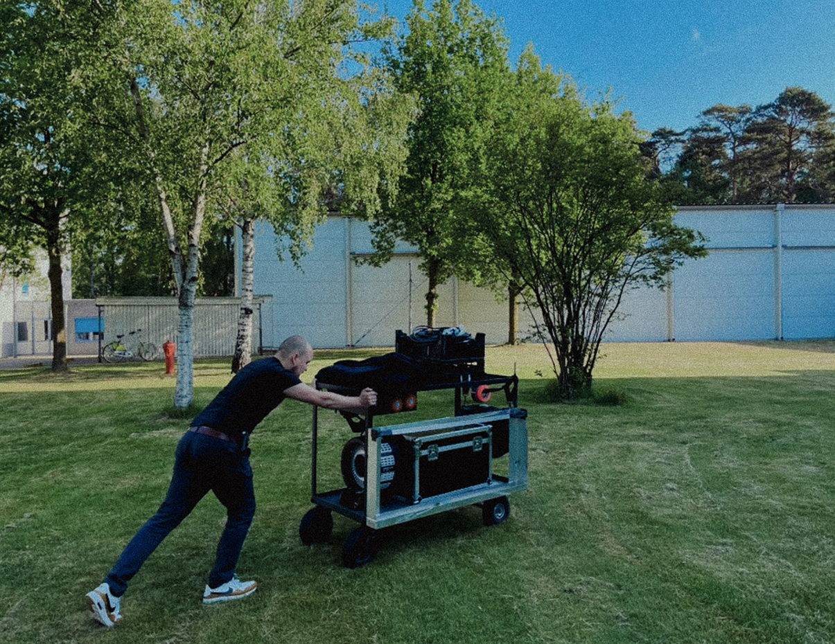 Man duwt een mobiele audio- of video-installatie op een grasveld met groene bomen en een witte muur op de achtergrond.