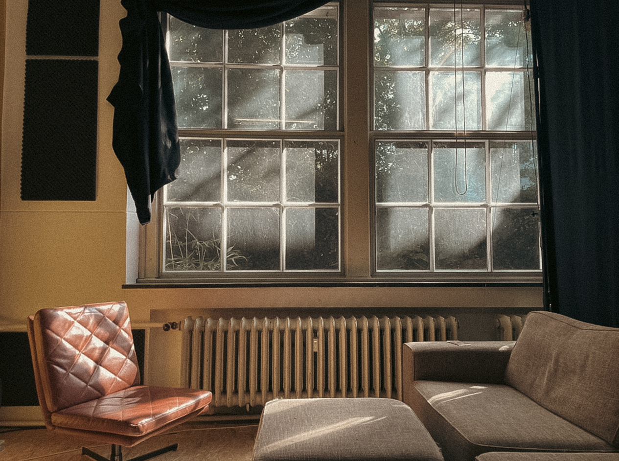 Sunlight streams through a large window illuminating a cozy living room with a pink leather chair, a beige sofa, a radiator beneath the window, and dark curtains.