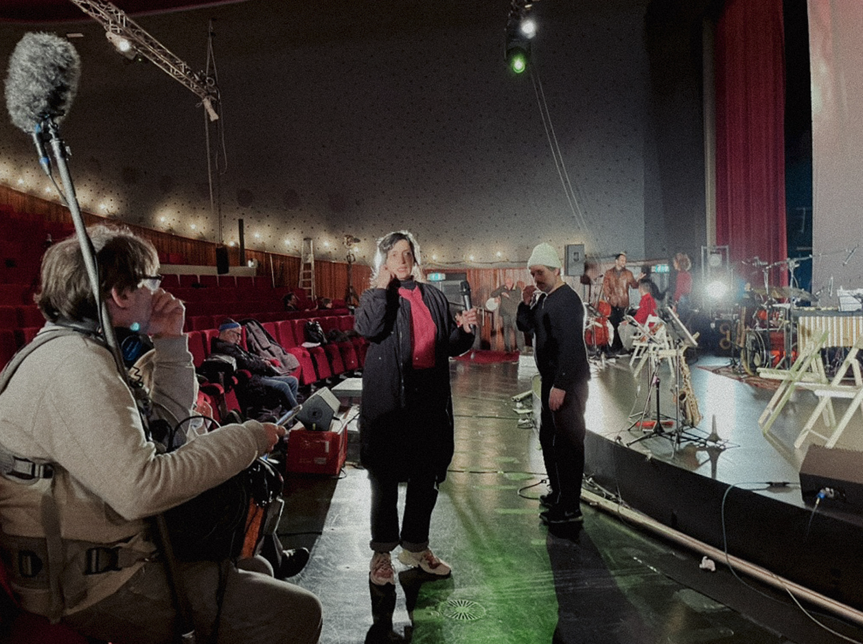 Een repetitieruimte of theater met mensen die zich voorbereiden op een uitvoering, met een camera en geluidsapparatuur.