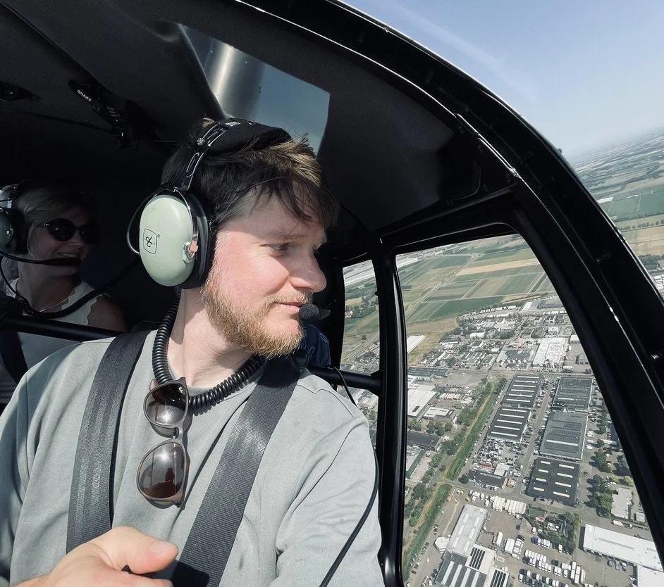 A man wearing a headset and sunglasses inside a helicopter cockpit, looking out at the landscape below, with a woman in the background also wearing a headset.