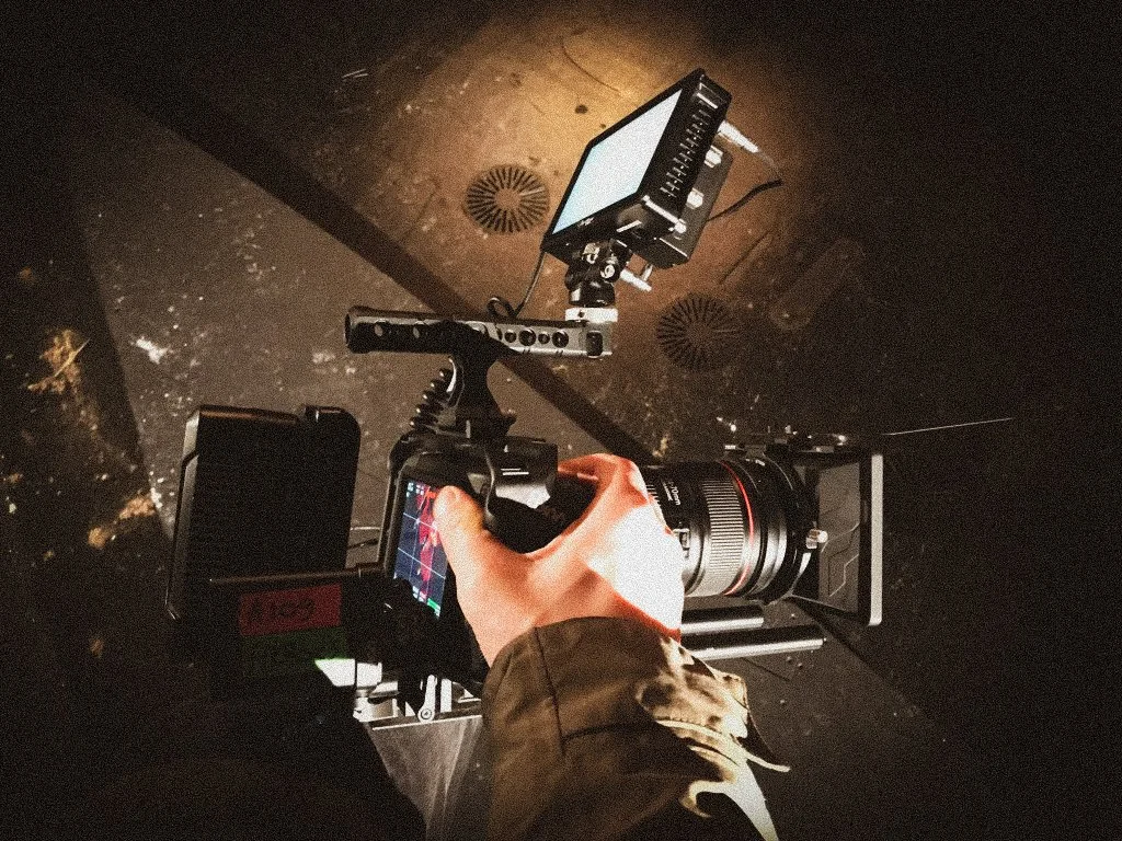 The image shows a camera mounted on a stabilizer, with a person's hand operating it. The camera is aimed at a computer monitor mounted on a rig, with a dark background.