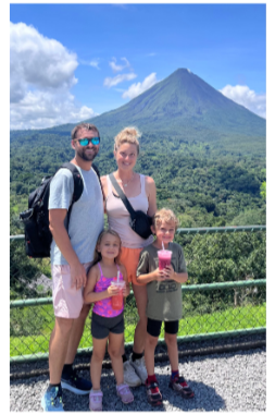 A family of four standing outdoors with a mountain and volcano in the background. The man has a beard and sunglasses, the woman has blonde hair, and two children are holding pink drinks. All are smiling.
