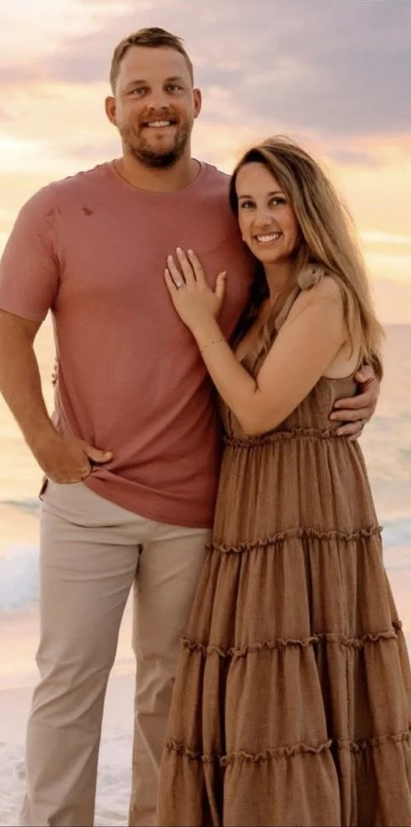 A man and woman smiling at the camera on a beach at sunset, with the woman showing her ring.