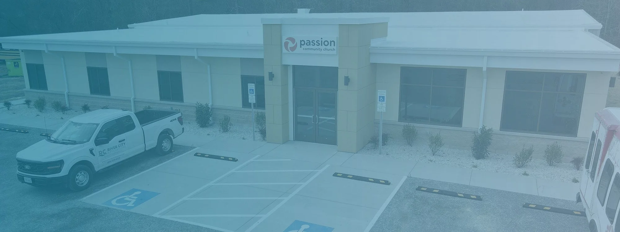 Exterior of Passion Community Church with a parking lot featuring two accessible spots, a white truck parked, and an ambulance nearby. The church has large windows and a sign above the entrance.
