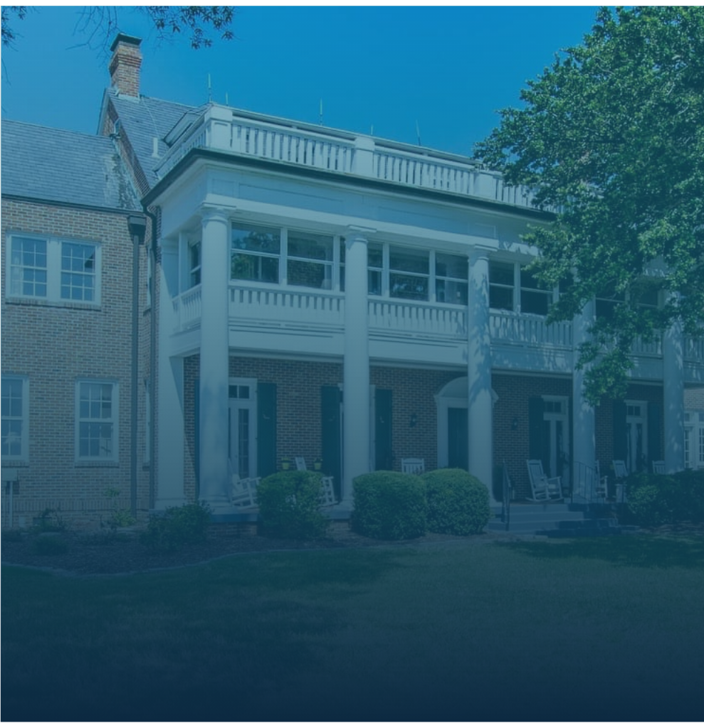 A large brick house with a porch supported by white columns, multiple windows, and a balcony on the upper level. There are trees and bushes in the front yard.