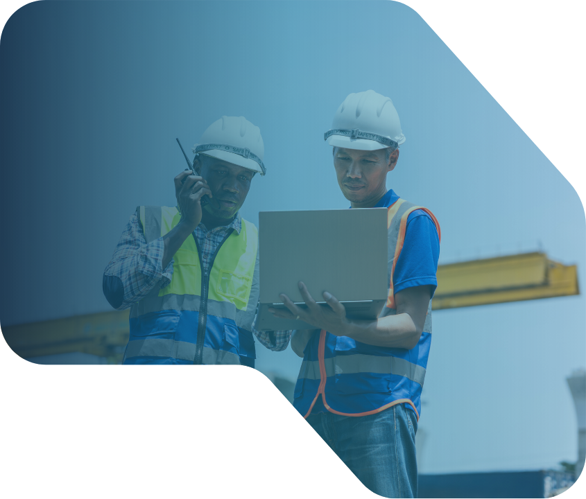 Two construction workers in safety vests and helmets look at a laptop at a construction site with a crane in the background.