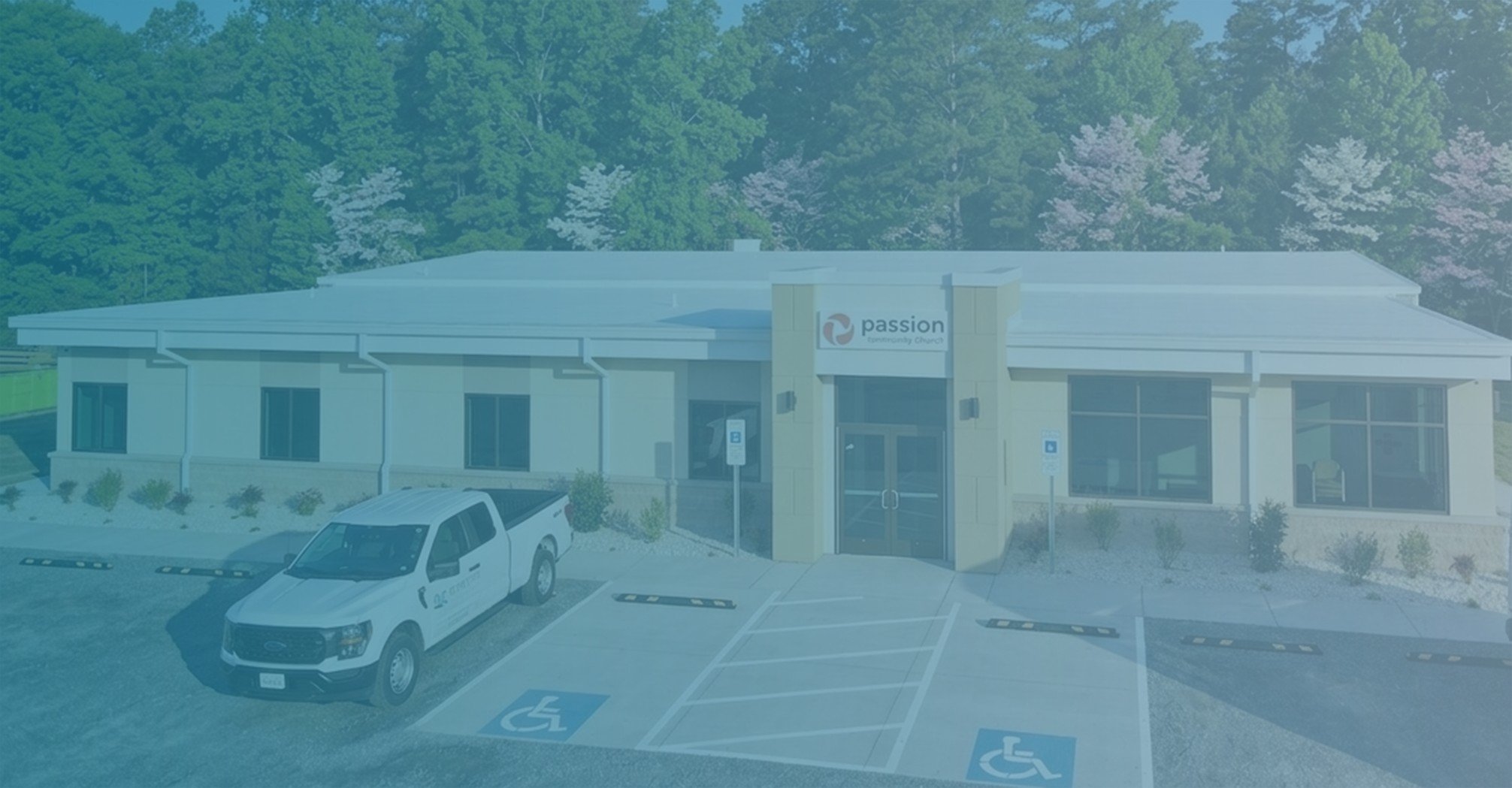 A single-story building with a sign that reads 'passion facingworld change' on the front. There are multiple parking spaces in front of the building, including designated handicap spots with blue markings. A white pickup truck is parked in the lot, and a forested area with trees is visible behind the building.