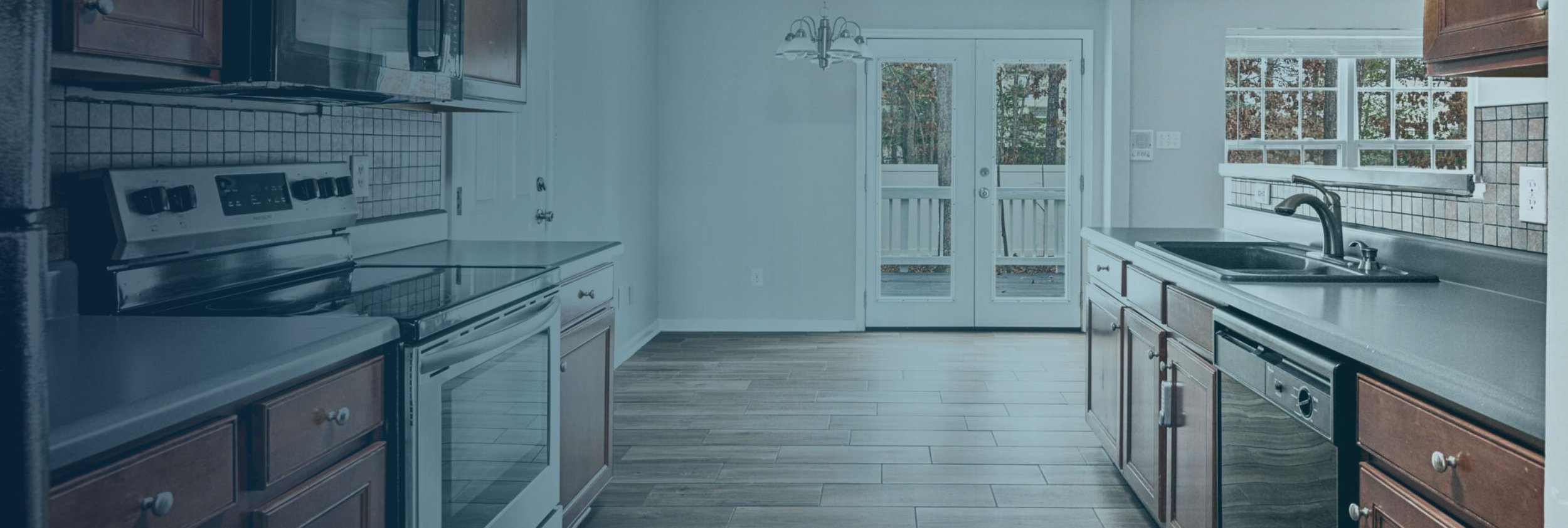 Empty kitchen with wooden cabinets, white countertops, black sink, and large glass doors leading to a deck with trees outside.