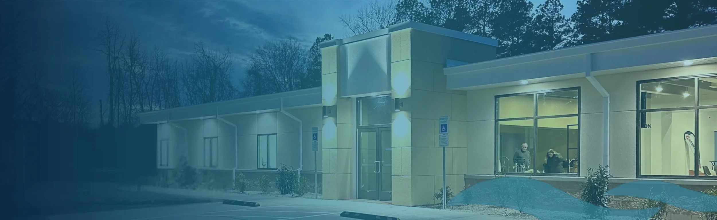 Exterior view of a modern building with large glass windows, nighttime, with interior lights on and people visible inside.