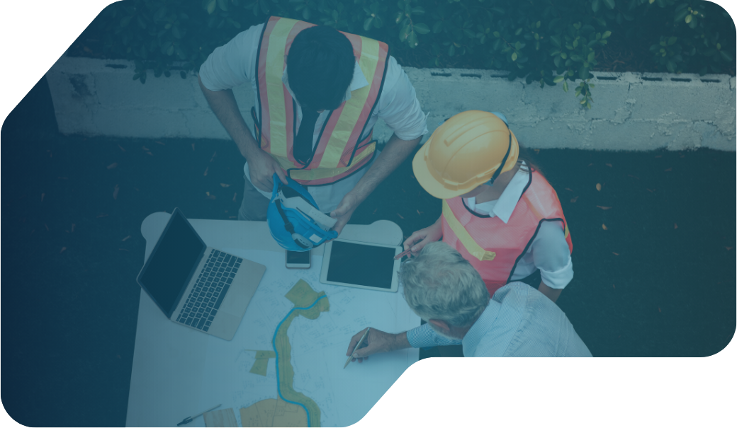 Overhead view of two engineers and a supervisor reviewing construction plans and digital devices at a table outdoors.