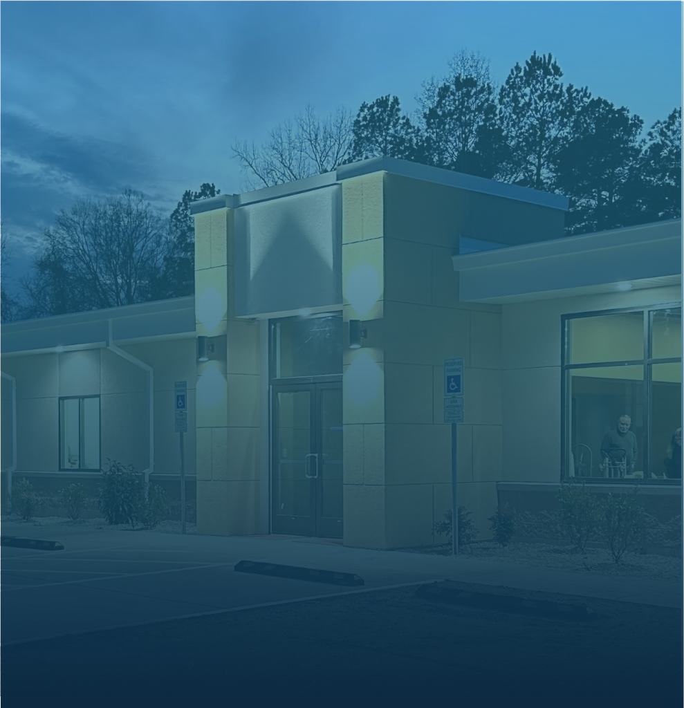Night view of a modern building with blue sky and trees in the background, exterior lights illuminating the entrance, with people visible inside through large windows, and a parking lot with handicapped parking signs outside.