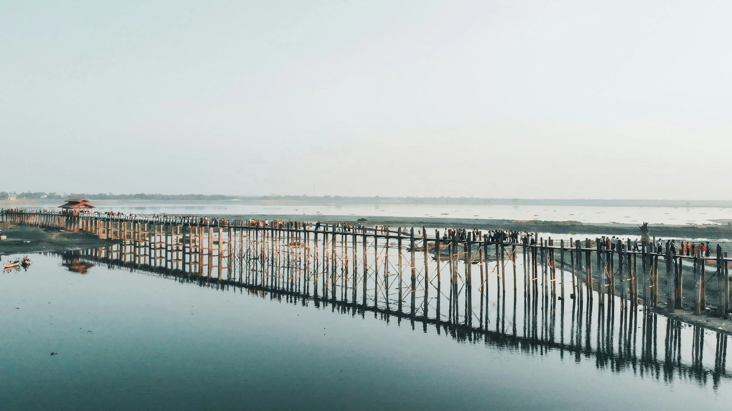 A long wooden pier extends over a calm body of water, with many people walking along it. The water reflects the pier and the sky, creating a mirror-like effect. In the distance, a small structure is at the end of the pier, and the landscape is flat with a clear sky.